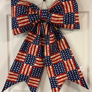 May include: A large fabric bow featuring a patchwork pattern of American flag motifs with blue fields of white stars and red and white stripes. The fabric has a distressed, vintage-style texture. The bow has two long, pointed tails hanging down.