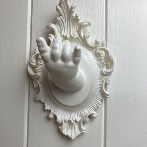 Puede incluir: Gancho de cerámica blanca con forma de mano con un marco blanco adornado. El gancho está fijado a una pared blanca.