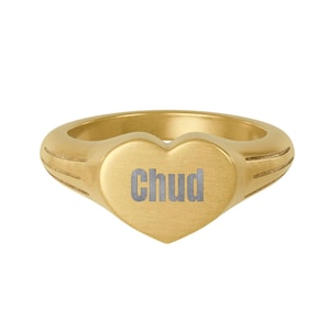 May include: A gold-colored ring with a heart-shaped face. The word "Chud" is engraved in silver on the heart. The ring has a smooth, polished finish and a band with etched details on each side.
