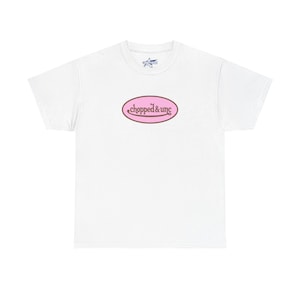 May include: White t-shirt featuring a pink oval design with the words "chopped & unc" in a brown font. The shirt has a crew neck and short sleeves. A small logo is visible near the neckline.