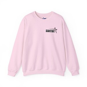 May include: A light pink crew neck sweatshirt with long sleeves. The sweatshirt features the text "SDIYBT" and a star graphic in black on the upper left chest area. The garment is made of a soft material and is suitable for casual wear.