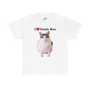 May include: White t-shirt featuring a graphic of a cat with brown and white markings. Above the cat is the text "I ❤️ Uncle Bao" in black, with a red heart symbol. The shirt has a crew neck.