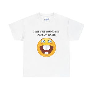 May include: White t-shirt with the text "I AM THE YOUNGEST PERSON EVER!" above a yellow emoji with a wide grin, blue eyes, and large white teeth. The shirt has a crew neck and short sleeves.