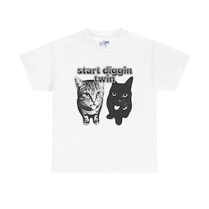 May include: White t-shirt featuring a graphic of two cats, one tabby and one black, with the text "start diggin twin" above them. The cats are facing forward with their tongues out. The shirt is made of a soft material.
