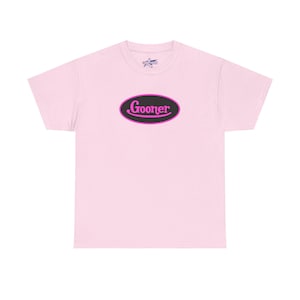May include: A light pink t-shirt with a crew neck. The shirt features a black oval with a pink border and the word "Gooner" in pink script. A small star logo is visible near the neckline.