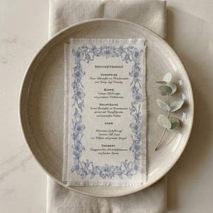 Könnte beinhalten: Eine Hochzeitsmenükarte mit blauem Blumenrand und Text auf Deutsch, auf einem beigen Teller mit einer Leinen-Serviette. Das Menü umfasst Gänge wie Suppe und Dessert. Ein Zweig Grün liegt auf dem Teller.