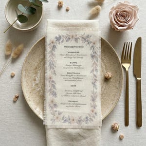 Könnte beinhalten: Eine Hochzeitsmenükarte auf einer Leinen-Serviette, verziert mit floralen und blattartigen Akzenten in sanften Pastellfarben. Die Serviette liegt auf einem beigen Teller, mit goldenem Besteck und einer rosa Rose. Trockenblumen und Grünzeug vervollständigen die elegante Tischdekoration.