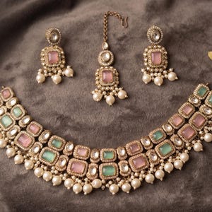 Könnte beinhalten: Ein Schmuckset bestehend aus einer Halskette, zwei Ohrringen und einem Maang Tikka. Die Stücke sind aus goldfarbenem Metall gefertigt und mit quadratischen rosafarbenen und mintgrünen Steinen, Kristallen und weißen Perlen verziert. Text: Exquisite Indian Kundan Bridal Set.