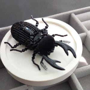 May include: A black beaded beetle brooch with large, curved mandibles. The insect-shaped accessory features a velvet-like texture and is embellished with black gemstones. The brooch is displayed on a white circular base.