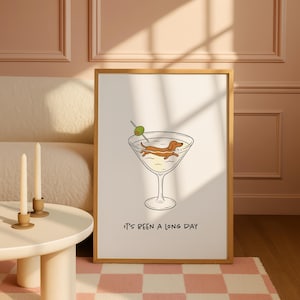 Może przedstawiać: Oprawiony wydruk z kresk&oacute;wkową ilustracją jamnika relaksującego się w kieliszku do martini z oliwką. Pod spodem wydrukowano tekst "It's been a long day". Grafika umieszczona jest na kremowym tle.