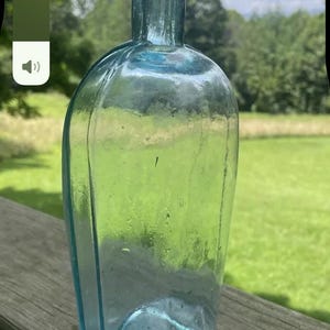 May include: A vintage, light blue glass bottle with a rounded body and a long neck. The bottle is empty and appears to be antique, with a slightly irregular shape. The background shows a green lawn and trees.