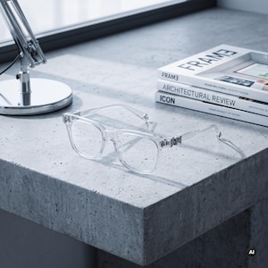 Puede incluir: Gafas transparentes con un diseño de montura cuadrada y detalles decorativos metálicos en las bisagras. Las gafas presentan una estructura ligera con patillas finas y lentes claras, colocadas sobre una superficie gris texturizada.
