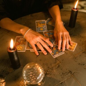 May include: A collection of tarot cards are arranged on a dark, textured surface. Two black candles with lit flames flank the cards. A crystal ball sits in the foreground. The scene is illuminated by the candle flames.