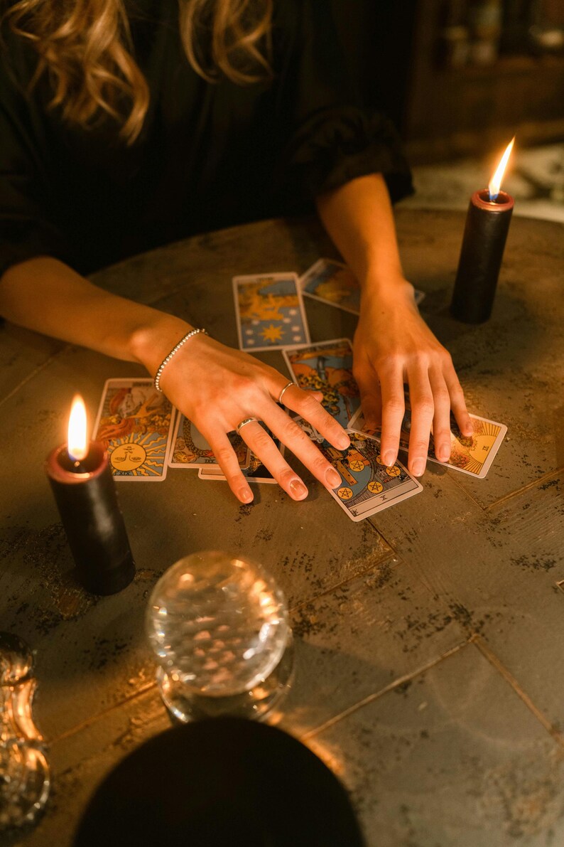 May include: A collection of tarot cards are arranged on a dark, textured surface. Two black candles with lit flames flank the cards. A crystal ball sits in the foreground. The scene is illuminated by the candle flames.