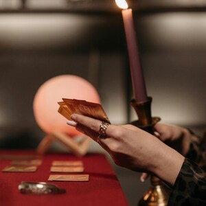 May include: A lit purple candle in a gold candlestick holder. A hand holds a deck of gold cards, with other cards on a red surface. A glowing orb and a decorative bracelet are also visible.