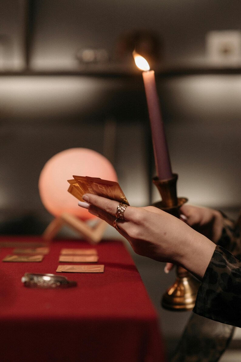 May include: A lit purple candle in a gold candlestick holder. A hand holds a deck of gold cards, with other cards on a red surface. A glowing orb and a decorative bracelet are also visible.
