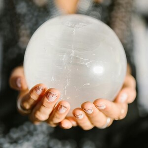 Pode incluir: Uma grande bola de cristal transparente &eacute; segurada em m&atilde;os em concha. A esfera tem uma superf&iacute;cie lisa e polida com veios internos sutis. As m&atilde;os t&ecirc;m unhas cuidadas com um design rosa claro e dourado. O fundo est&aacute; desfocado.