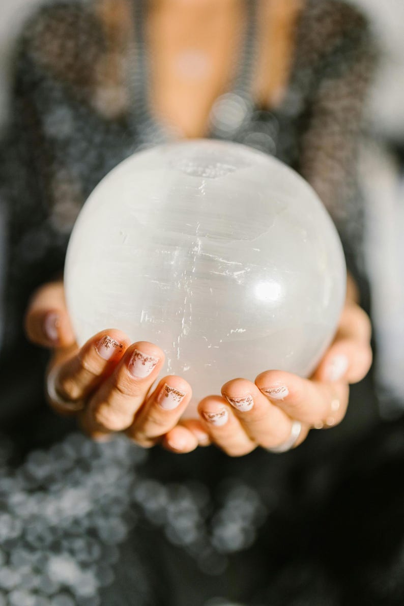 Pode incluir: Uma grande bola de cristal transparente &eacute; segurada em m&atilde;os em concha. A esfera tem uma superf&iacute;cie lisa e polida com veios internos sutis. As m&atilde;os t&ecirc;m unhas cuidadas com um design rosa claro e dourado. O fundo est&aacute; desfocado.
