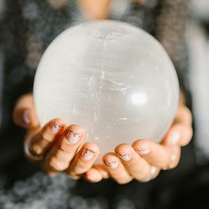 May include: A large, clear crystal ball is held in cupped hands. The sphere is smooth and translucent, with subtle internal veining. The hands have manicured nails with a gold glitter design. The background is blurred, suggesting a focus on the crystal ball.