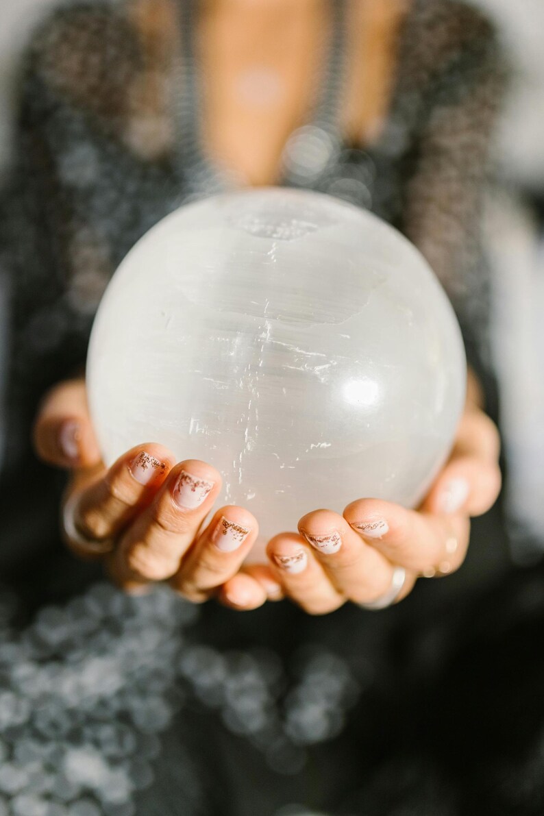 May include: A large, clear crystal ball is held in cupped hands. The sphere is smooth and translucent, with subtle internal veining. The hands have manicured nails with a gold glitter design. The background is blurred, suggesting a focus on the crystal ball.