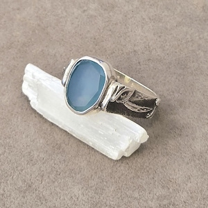 May include: A silver ring featuring a faceted, oval-shaped light blue stone set in a bezel. The metal band displays a textured, dark oxidised finish with a carved leaf or feather motif on the side, resting on a white, translucent crystal shard.