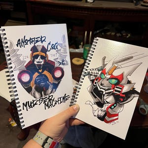 May include: Two spiral-bound sketchbooks with robot illustrations. One has a robot design and the text "ANOTHER COG IN THE MURDER MACHINE." The other shows a robot with red and white details. The sketchbooks are held in a person's hand.