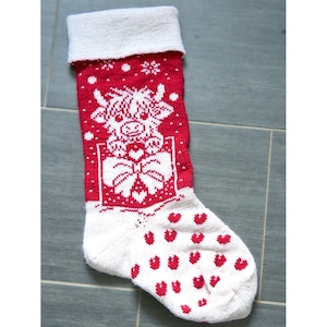 Op de afbeelding: Een rood-witte gebreide kerstsok met een centrale afbeelding van een Schotse hooglander die in een cadeauverpakking met een grote strik zit. De boord is effen wit en het voetgedeelte is wit met een patroon van kleine rode hartjes overal.