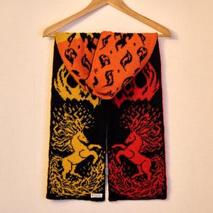 May include: A knitted scarf with a hood, featuring a black base with yellow and red unicorn designs. The hood and top section of the scarf are orange with black leaf patterns. The scarf is hanging on a wooden hanger.