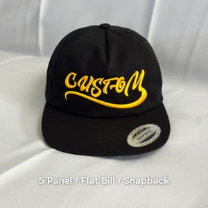 May include: Black snapback hat with a flat bill. The word "CUSTOM" is embroidered in yellow script across the front. The hat has a 5-panel design and a snapback closure. A small, silver tag is on the bill.
