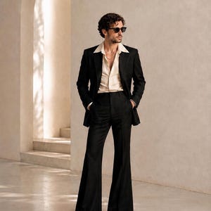 Può includere: Un completo composto da una giacca nera e pantaloni abbinati a vita alta con taglio a zampa, indossati con una camicia color crema abbottonata. L'abbigliamento include occhiali da sole scuri e scarpe eleganti nere, creando un look strutturato con pantaloni a gamba larga.
