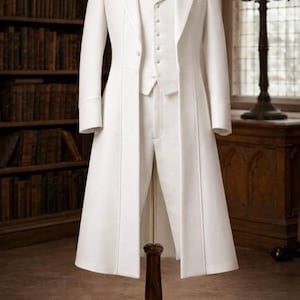 May include: A formal white suit displayed on a wooden mannequin. The suit includes a long coat, vest, and trousers. The coat has a long, flowing design. The suit is displayed in a room with a bookshelf and antique furniture.