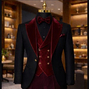 May include: A formal black suit with a burgundy velvet lapel, vest, and bow tie. The vest features gold buttons. The suit includes matching burgundy trousers and a black dress shirt. The suit is displayed on a mannequin.