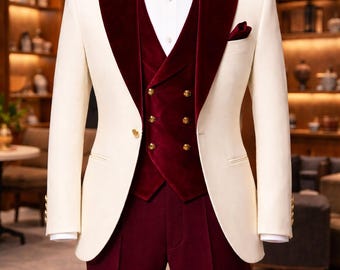 May include: A cream-coloured suit with a burgundy velvet lapel, waistcoat, and trousers. The waistcoat features gold buttons. A matching burgundy bow tie and pocket square complete the formal ensemble. The suit is displayed on a mannequin.