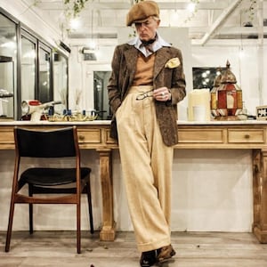 Puede incluir: Una persona con una chaqueta de tweed marr&oacute;n, pantalones color beige y una gorra marr&oacute;n. El atuendo incluye una camisa azul claro, una bufanda estampada y un pa&ntilde;uelo de bolsillo amarillo. La persona sostiene gafas cerca de una mesa de madera.