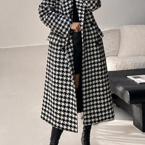Puede incluir: Un abrigo largo de pata de gallo en blanco y negro. El abrigo llega a la rodilla y tiene un corte entallado. La persona lleva botas negras. El abrigo es una prenda de abrigo clásica y elegante.