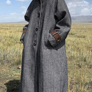 May include: A grey herringbone overcoat with a high collar and brown detailing on the collar and cuffs. The coat features large pockets and button fastenings. The person is standing in a field.