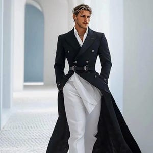 May include: A long, black double-breasted overcoat, paired with a white shirt and trousers, and a black belt. The outfit is completed with black shoes. The image is taken in a white hallway. The coat's length is a key feature.