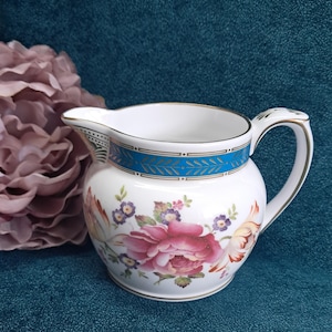 May include: A white porcelain cream jug featuring a vibrant floral pattern with large pink roses and smaller purple flowers. The rim is decorated with a bright blue band accented by gold trim, which also outlines the handle and the pouring spout.