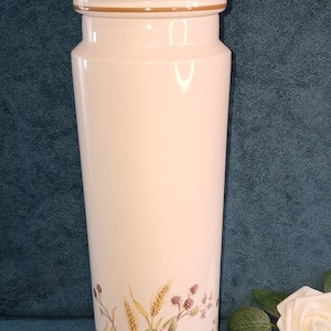 May include: A tall, cream-coloured ceramic jar with a lid and a decorative band. The jar features a floral design with wheat, berries, and leaves in shades of brown, green, and red. The jar has a gold-coloured trim.