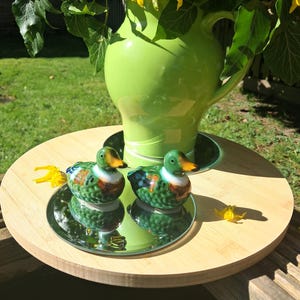 May include: A vibrant green ceramic jug displays yellow flowers and green foliage. Two duck ornaments, featuring green, white, and brown patterns, are placed on mirrored saucers on a wooden tray. The scene is set outdoors.