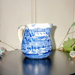 May include: Small blue and white ceramic jug with a handle. The jug features a scenic landscape design with a bridge and trees. The design is in a classic blue and white colour scheme, reminiscent of traditional pottery.