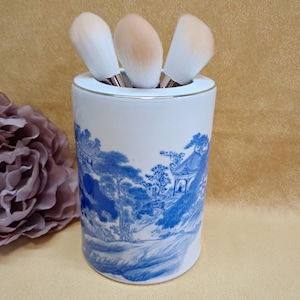 May include: A cylindrical white ceramic holder featuring a blue landscape pattern with trees and buildings. Three makeup brushes with white and tan bristles are placed inside the holder. The rim of the holder has a thin metallic silver trim.