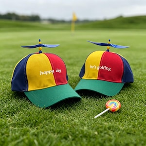 Może przedstawiać: Dwie kolorowe czapki golfowe z śmigłami na górze, jedna z napisem "happy day", a druga "he's golfing". Czapki mają zielony daszek, żółto-czerwoną koronę i granatowy bok. Tęczowy lizak leży na trawie.