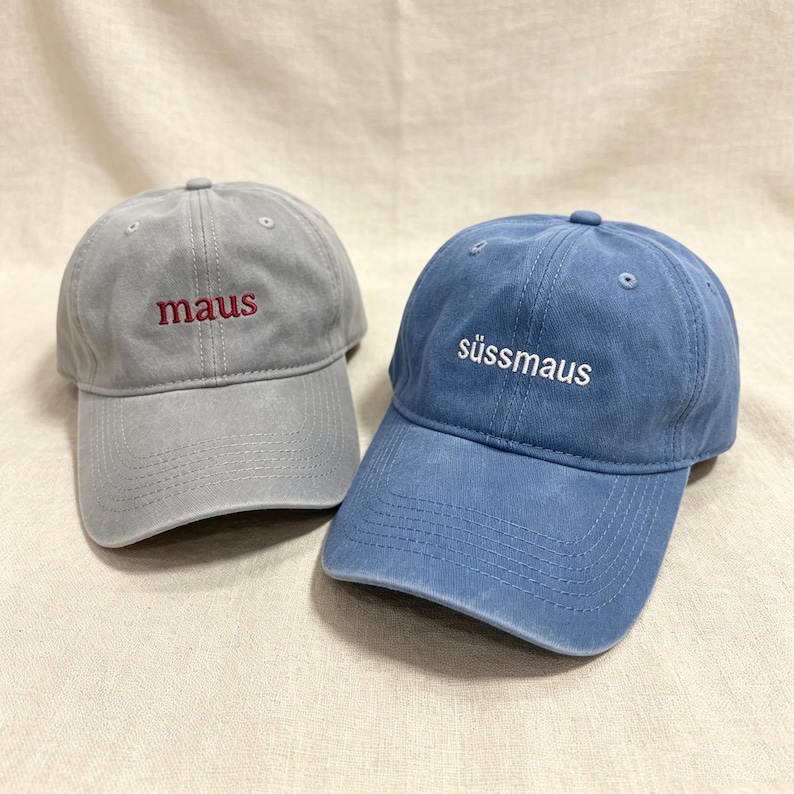 May include: Two baseball caps with embroidered text. The left cap is light gray with the word "maus" in red thread. The right cap is blue with the word "s&uuml;ssmaus" in white thread. Both caps feature a curved brim and a six-panel construction.