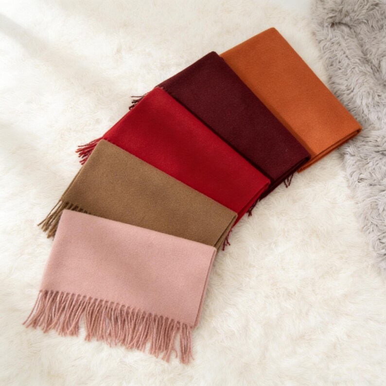 May include: A collection of six rectangular scarves in various colors, including pink, tan, red, burgundy, and orange. Each scarf has a fringed edge. The scarves are arranged on a white, fluffy surface.