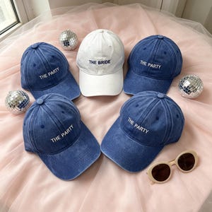 Könnte beinhalten: Vier blaue Baseballkappen aus Denim mit dem weißen gestickten Text THE PARTY und eine weiße Baseballkappe mit dem blauen gestickten Text THE BRIDE, begleitet von zwei kleinen silbernen Discokugeln und einer beigen, ovalen Sonnenbrille.