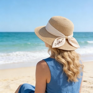 Peut inclure: Un chapeau de soleil en paille beige avec un large bord et un ruban crème noué en nœud. Le nom "Sophie" est écrit sur le nœud en cursive. Le chapeau est porté par une personne sur une plage avec l'océan en arrière-plan.