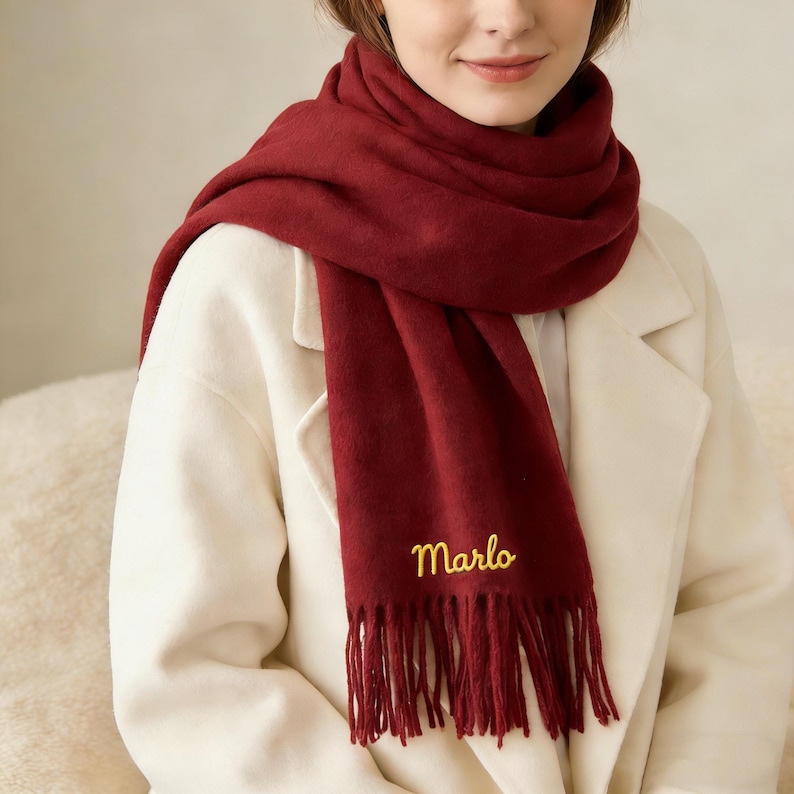 May include: A burgundy scarf with the name "Marlo" embroidered in gold thread. The scarf is fringed at the bottom and is draped over a white coat. The scarf appears soft and warm, suitable for cold weather.