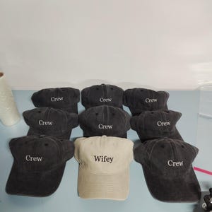 May include: A collection of eight baseball caps arranged in a group. Seven caps are dark charcoal gray with the word Crew embroidered in white thread. One cap is light beige with the word Wifey embroidered in black thread on the front panel.