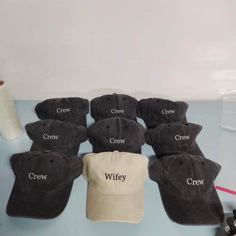 May include: A collection of eight baseball caps arranged in a group. Seven caps are dark charcoal gray with the word Crew embroidered in white thread. One cap is light beige with the word Wifey embroidered in black thread on the front panel.